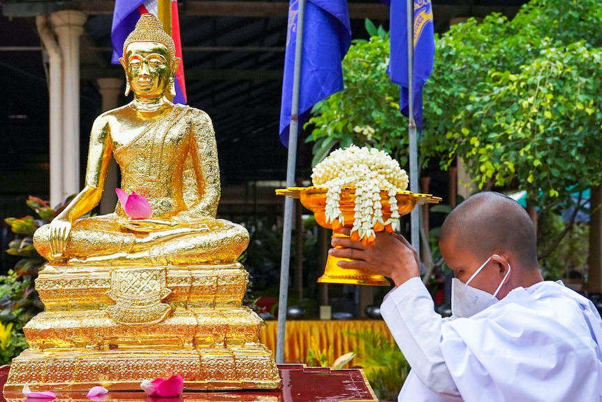 A Cambodian nun in a face mask holds a golden bowl filled with flowers next to a golden Buddha statue
