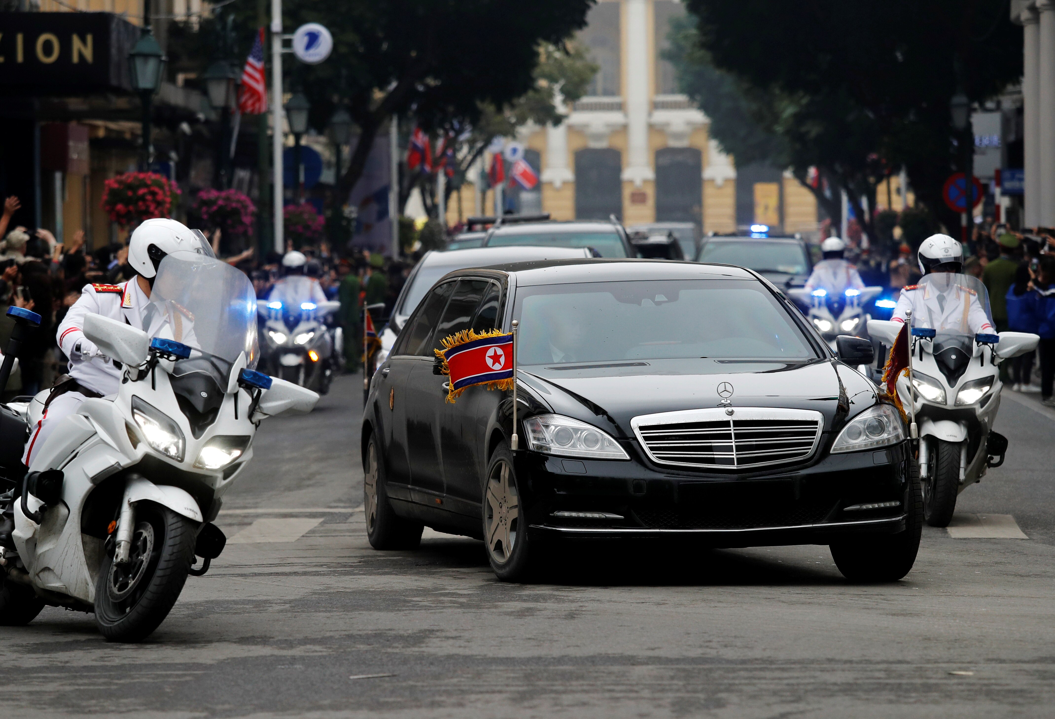 a limousine with North Korean flags on it is escorted by motorbikes and security officers.