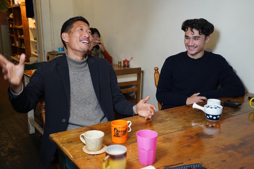 Two men sit at a table in a restaurant, one has his arms out and is laughing, the other looks on and smiles.