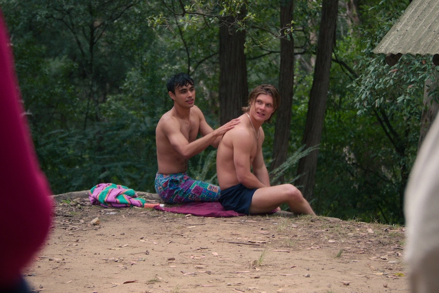 Against a bush backdrop, a shirtless teenage boy applied suncream to another's back. 