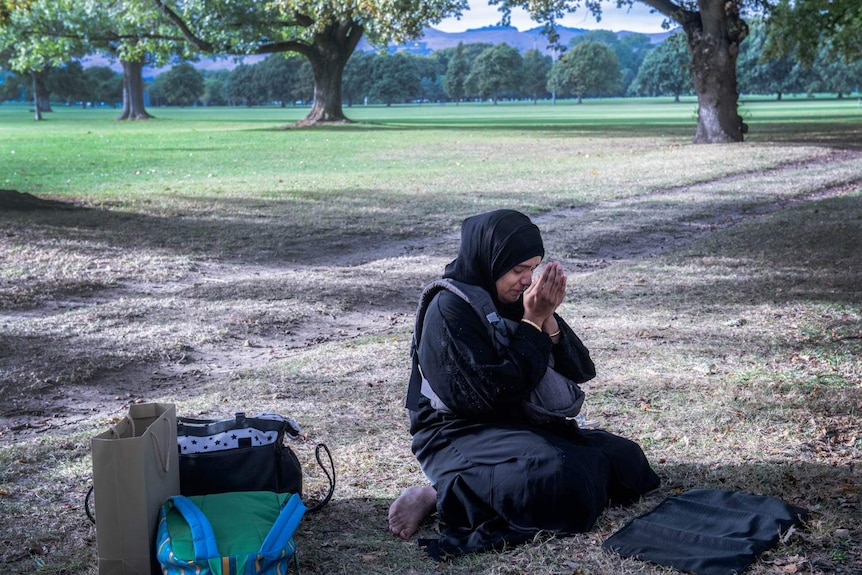 A woman in a hijab leans on her haunches in a park, holding her hands to her face