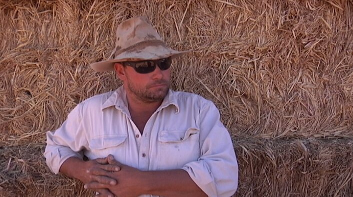 Man in hat and sunnies has a neutral face as he stands in front of a backdrop of hay