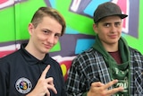 Two teenage boys make hand signals in front of a wall decorated with grafitti.