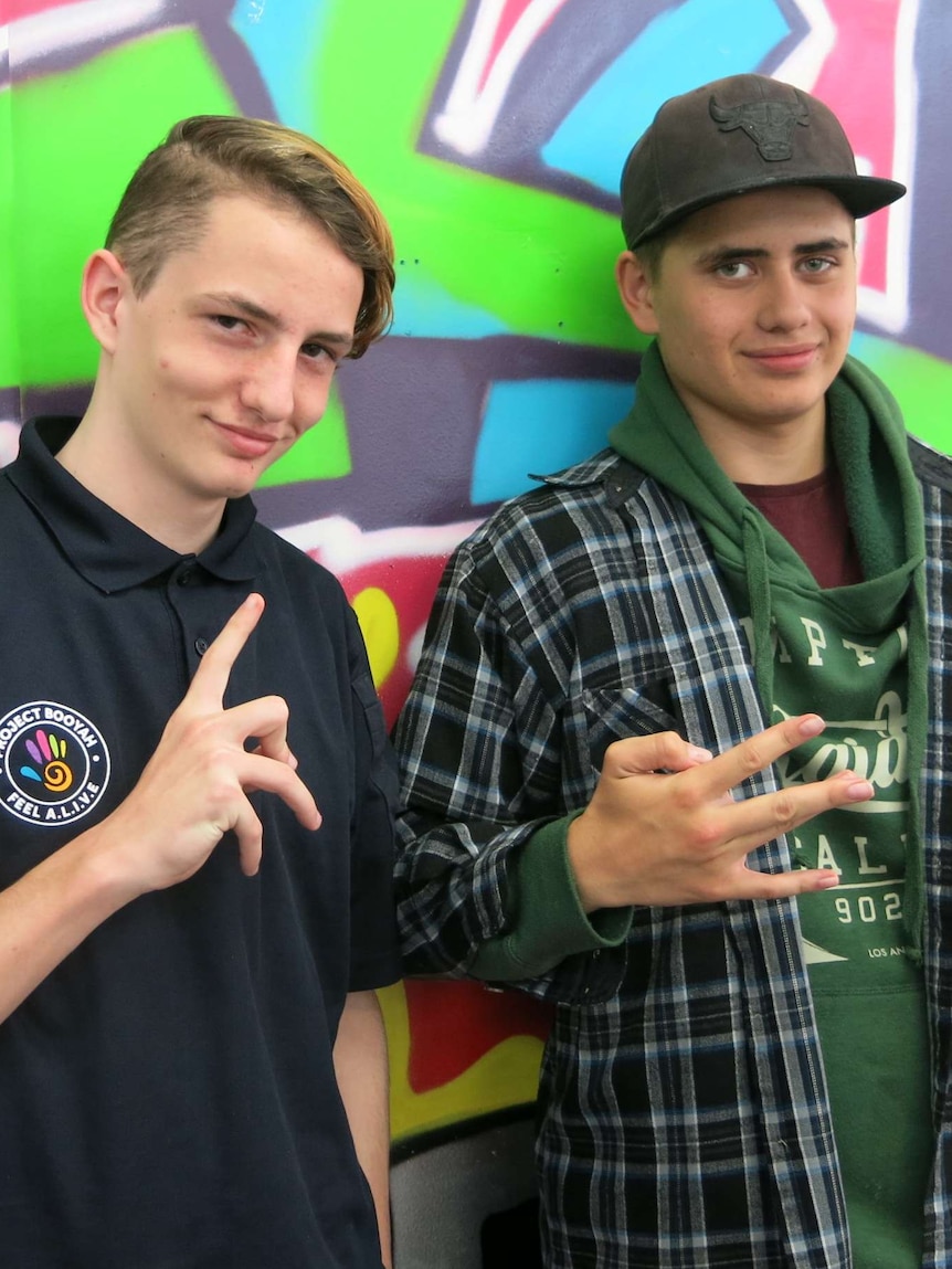 Two teenage boys make hand signals in front of a wall decorated with grafitti.