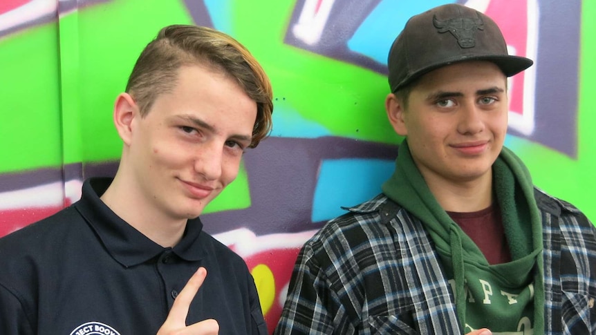 Two teenage boys make hand signals in front of a wall decorated with grafitti.