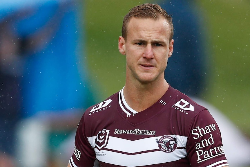 A Manly NRL player stands while holding a rugby league in his right hand.