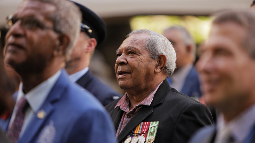 Indigenous man in a suit with medals watching an unveiling of a statue.