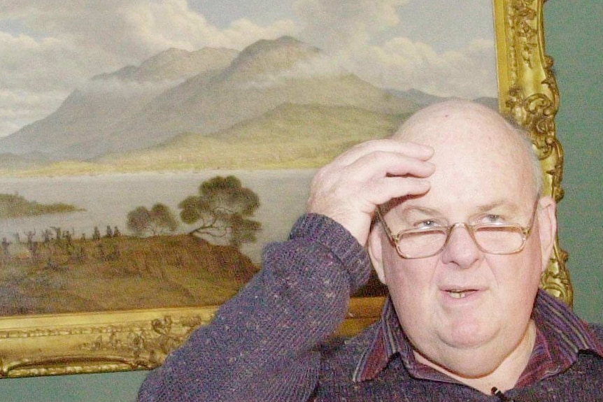 Les Murray stands with his hand on his head. He is in front of a landscape painting in a gold frame, and he is speaking.