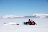 In a snowy environment, someone is pictured on a skidoo with a sleigh behind it.