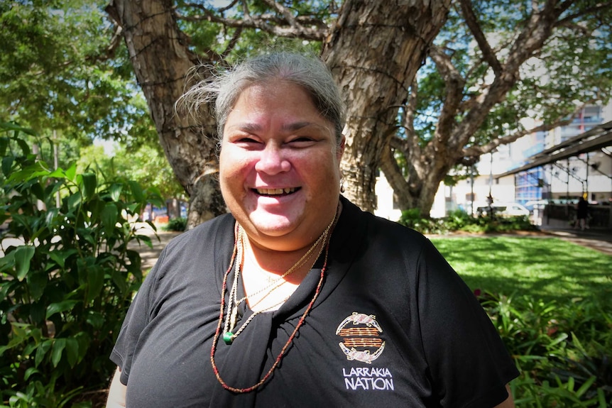 Woman smiling wearing a black Larrakia Nation shirt beside a tree in a city park. Sunny.