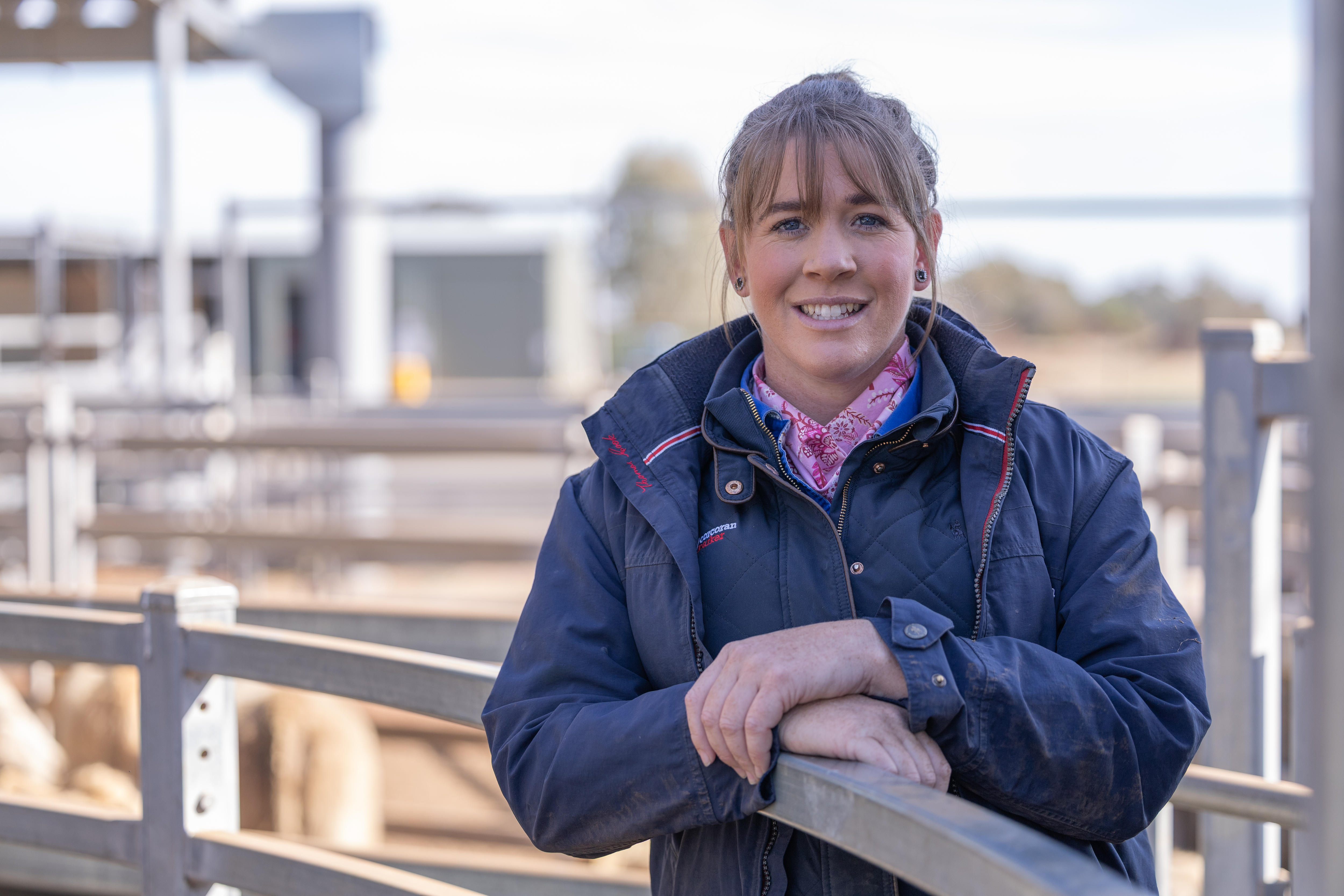 A woman in a shirt and coat smiles while leaning against the fence at a livestock yard