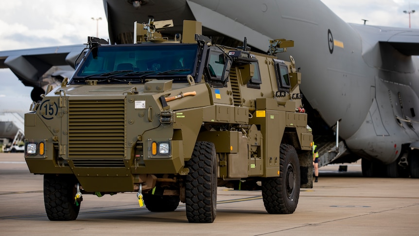 A Bushmaster sporting Ukrainian flags stands in front of a Globemaster airplane on the tarmac