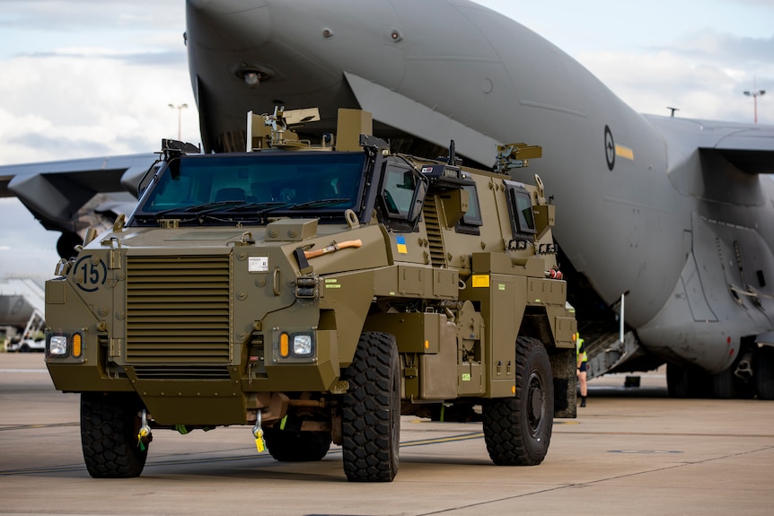 A Bushmaster sporting Ukrainian flags stands in front of a Globemaster airplane on the tarmac.