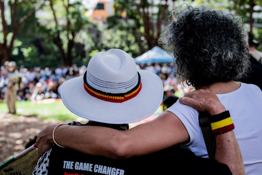 Two people with their arm around each other displaying indigenous colours on a wrist band and around a hat. 