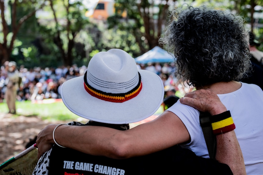 Two people with their arm around each other displaying indigenous colours on a wrist band and around a hat. 