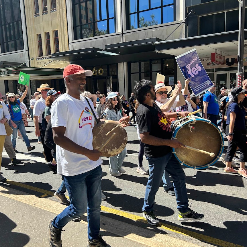 Supporters holding 'Yes' signs marching down the street, with two men in the front of the image holding drums.
