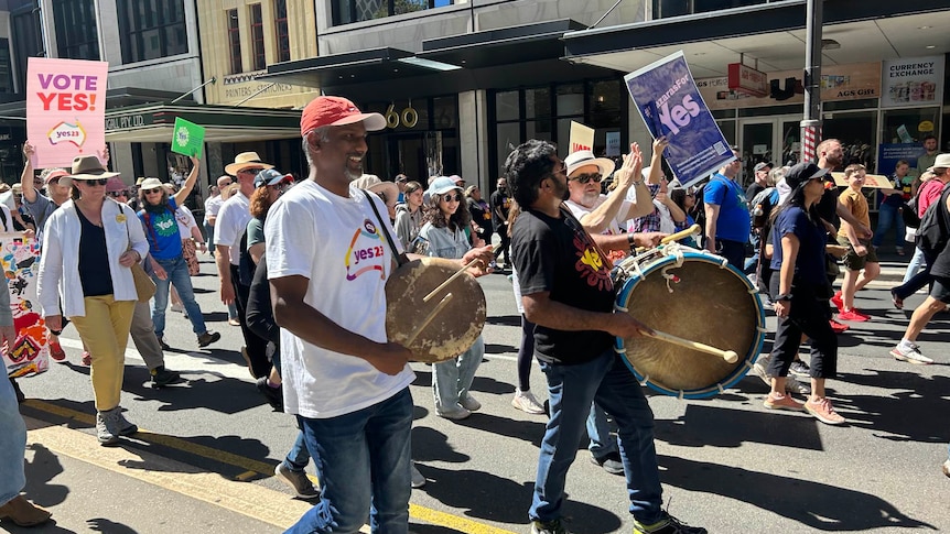 Supporters holding 'Yes' signs marching down the street, with two men in the front of the image holding drums.