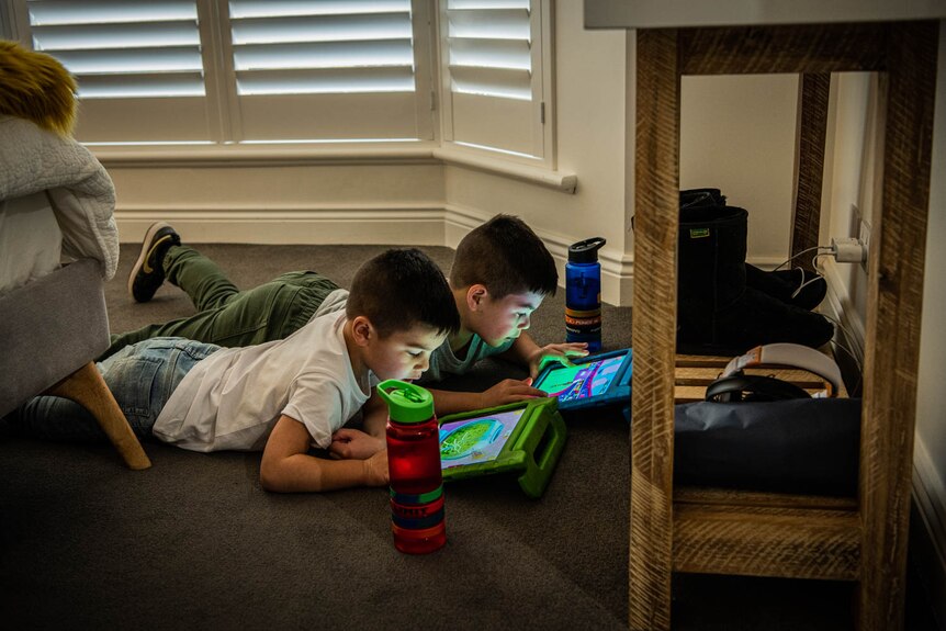 Two young boys lie on the floor playing on their ipads