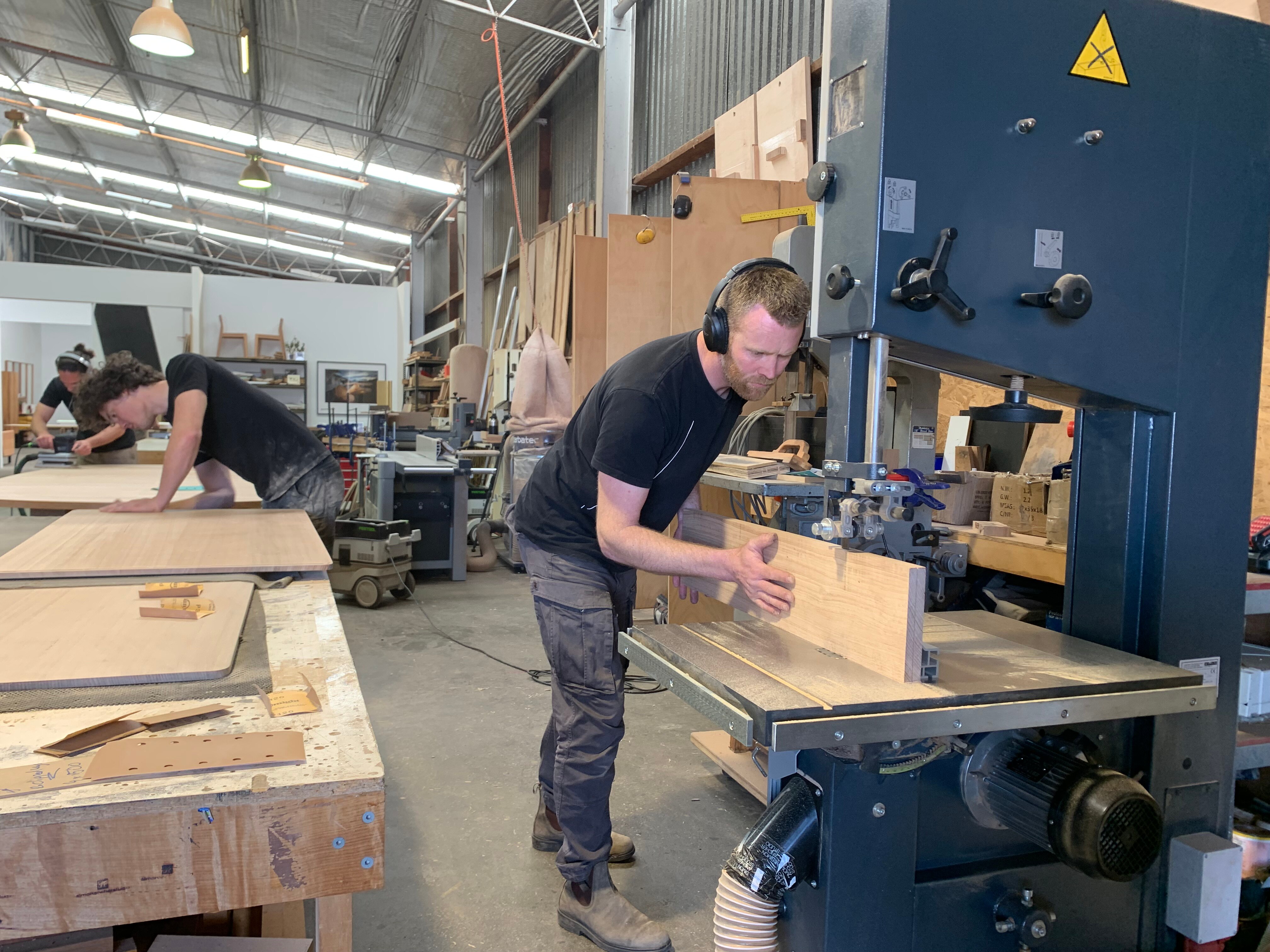 Man standing at heavy machinery in a workshop, pushing wood through a saw. 