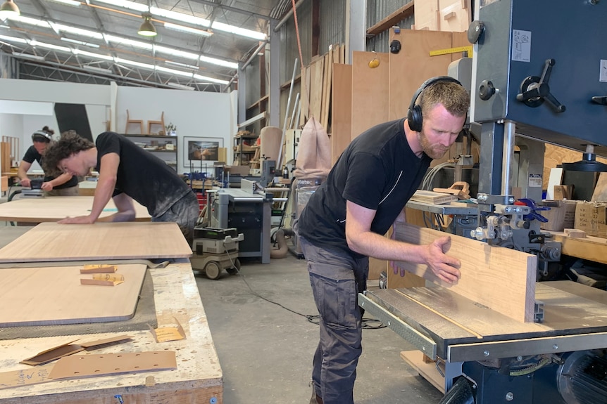 Man standing at heavy machinery in a workshop, pushing wood through a saw. 