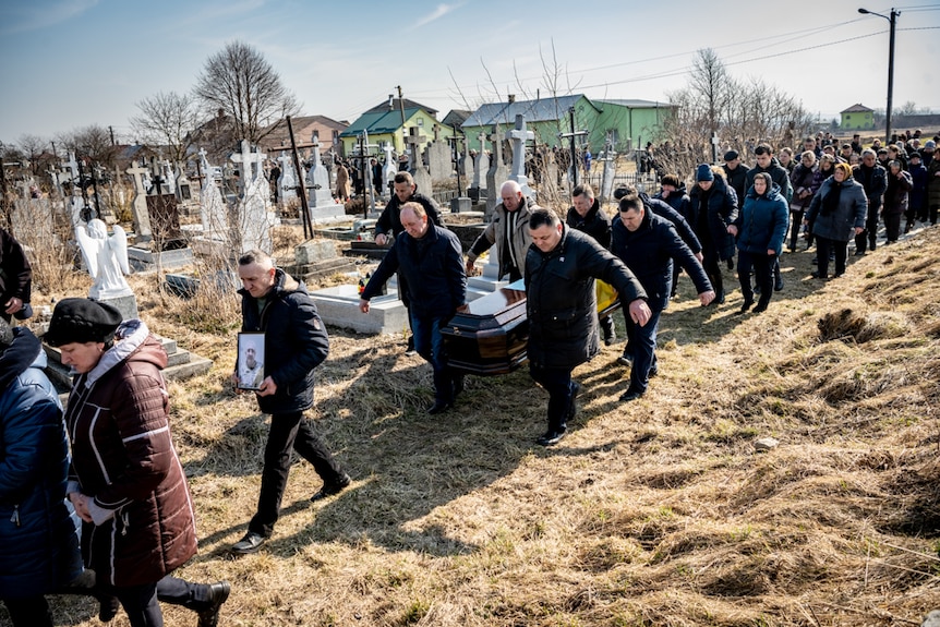 Men carry the coffin through the cemetery with villagers following behind.