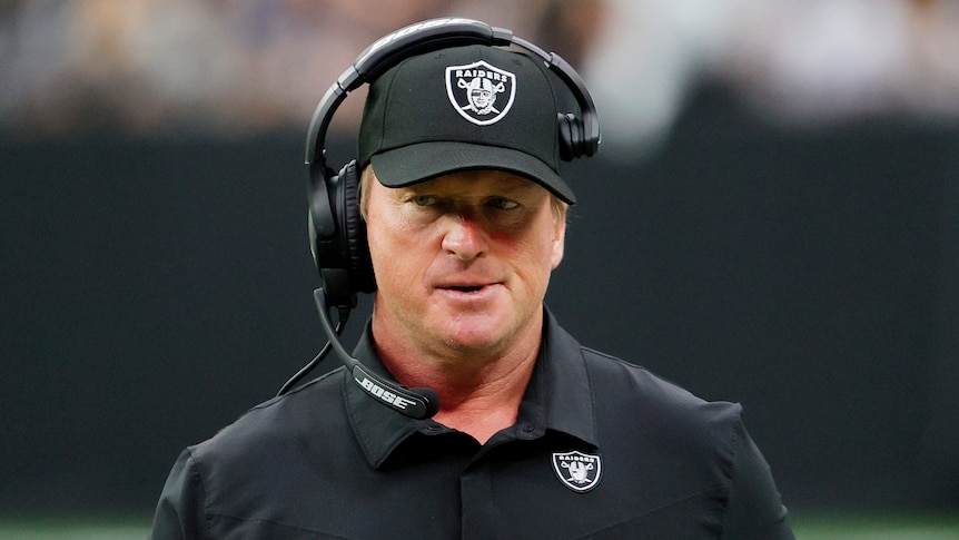 A coach wearing a black shirt and cap with the "Raiders" logo on it looks serious as he talks into a headset during an NFL game.