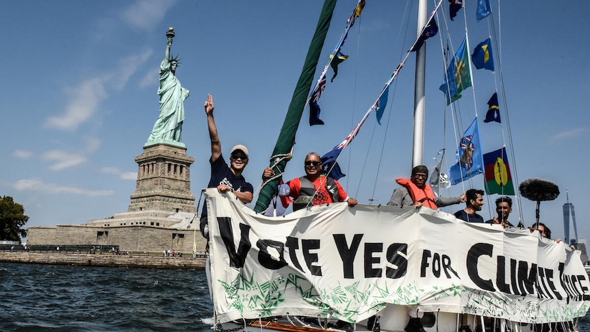 In front of the statue of liberty, a group are on a white boat holding up signs and Pacific flags.