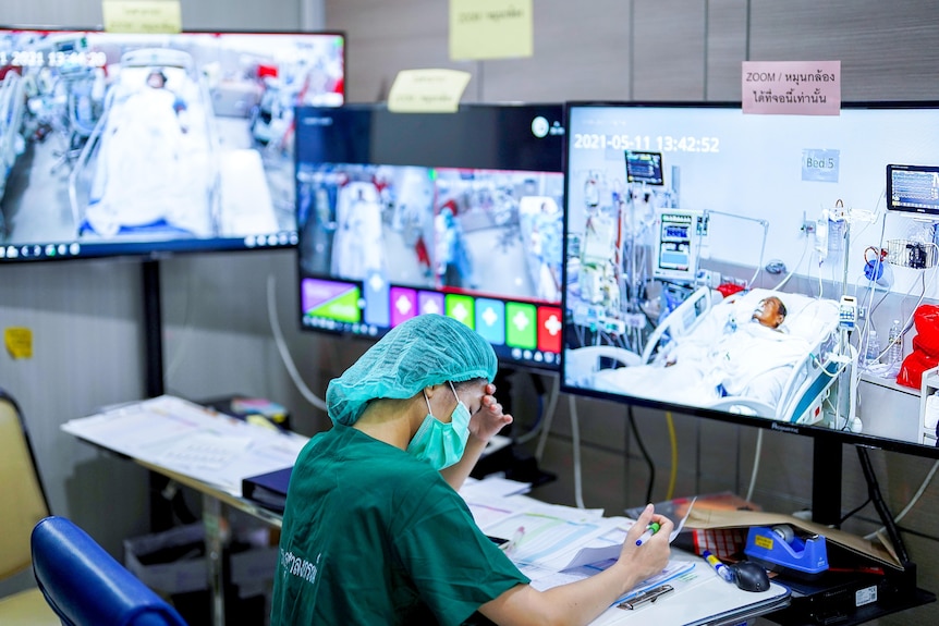 A woman in scrubs and a mask at a desk surrounded by screens showing patients in hospital beds