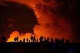 Silhouttes of people watching as large red and orange smoke clouds rise from the ground.
