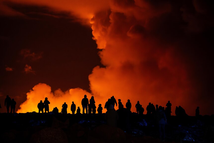 Silhouttes of people watching as large red and orange smoke clouds rise from the ground.