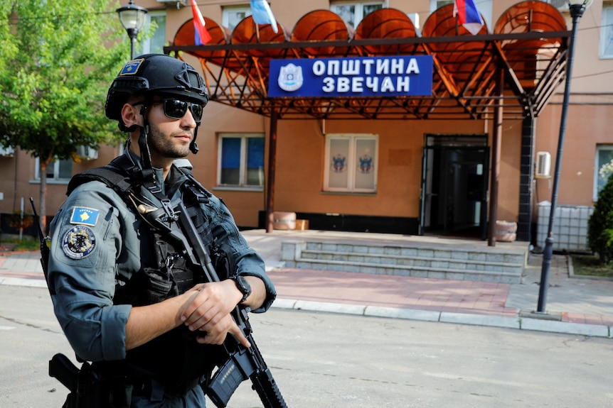 A young white man in a grey police uniform, with a helmet and combat vest and holding a rifle, stands guard outside a building.