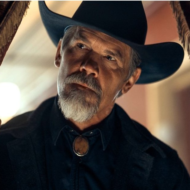 A man with a grey beard and black cowboy hat peers through a fringed curtain