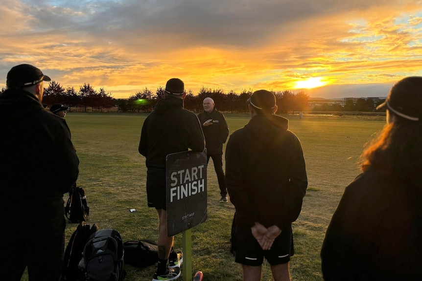 Rob de Castella speaking to a group of runners wearing black jumpers and caps with the setting sun behind them.