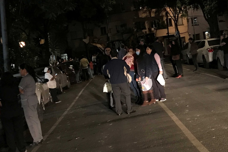 People stand on a street at night holding belongings.