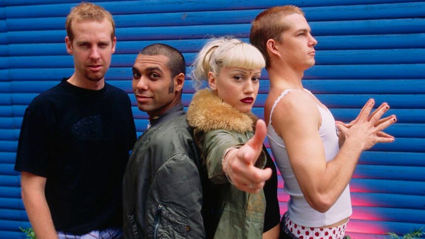 Three men and a woman from band No Doubt stand back to back before a blue wall. Gwen Stefani points fingers at camera.
