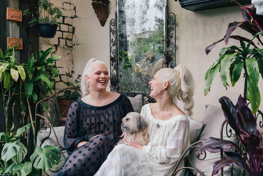 Two white women dressed in sheer dresses - one white, one black - and with matching platinum blonde hair sit laughing.