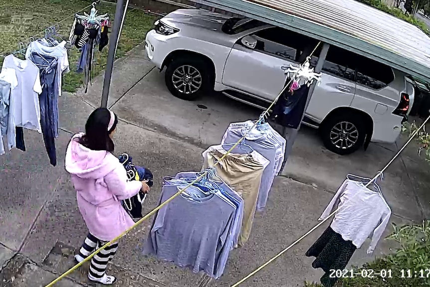 A woman wearesa pink dressing gown and black and white stripped pants. She carries a basket of washing to a clothes line