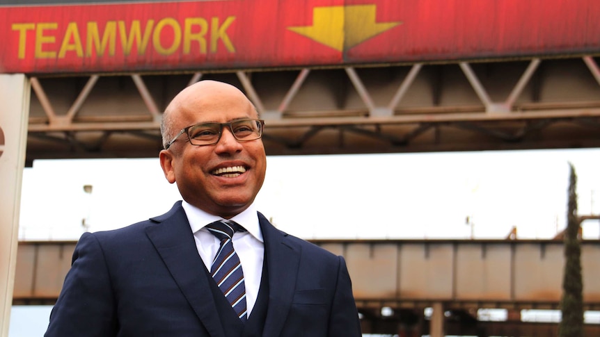 Sanjeev stands outside steelworks beneath a sign that says Teamwork.