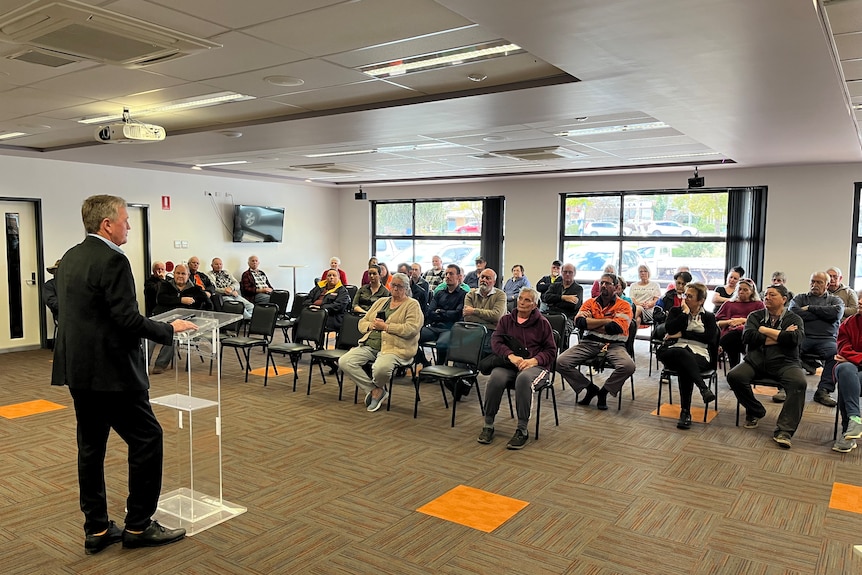 About 30 people sit inside a room listening to a person standing in front speaking to them 