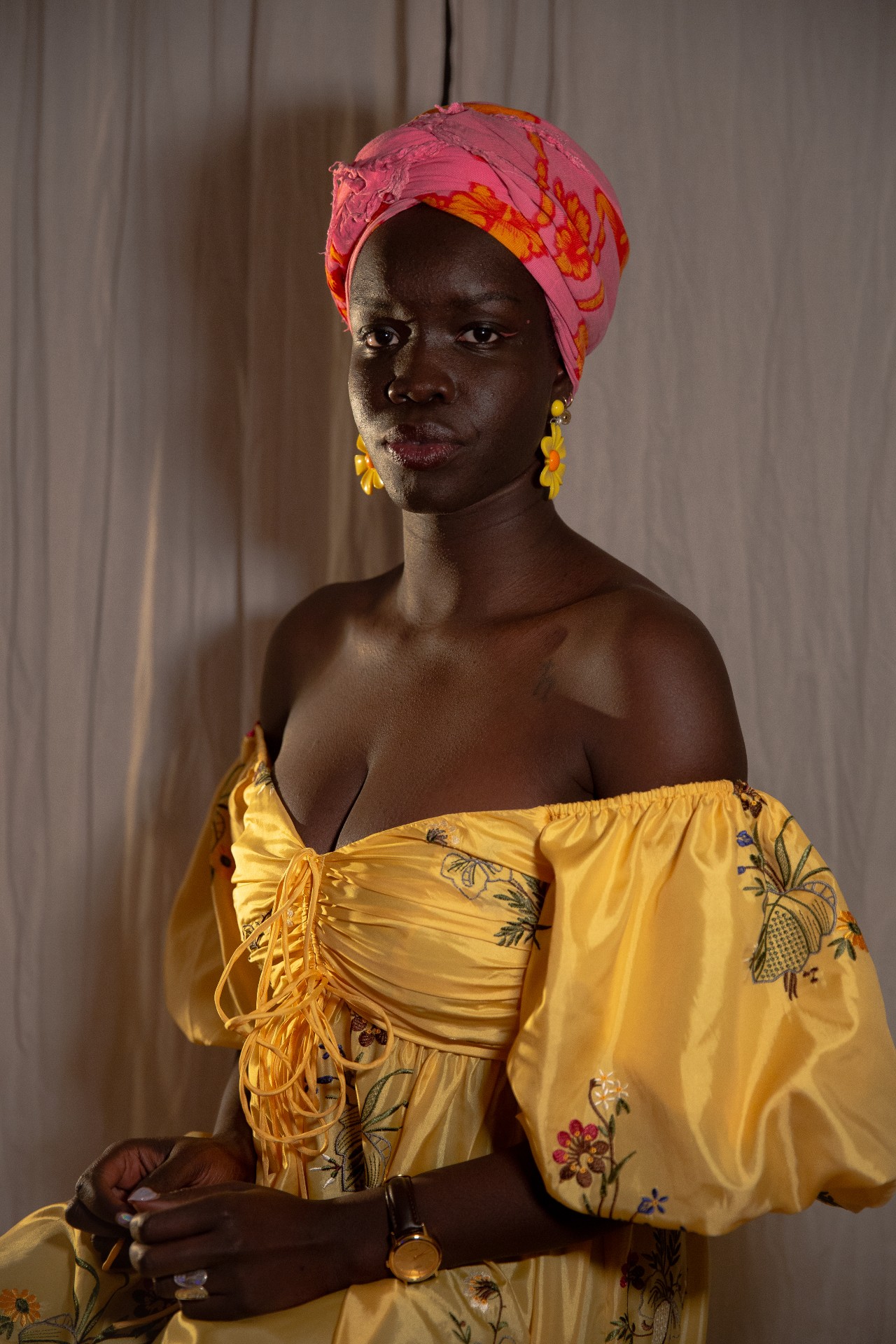 A 20-something African woman poses formally, her back straight, wearing a yellow dress and pink turban, her hands in her lap