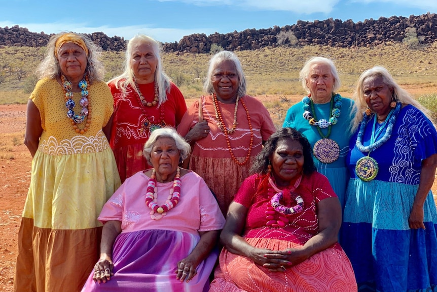 Community elders in colourful dresses pose for photo in front of desert with spinifex, wall of stacked large stones and blue sky