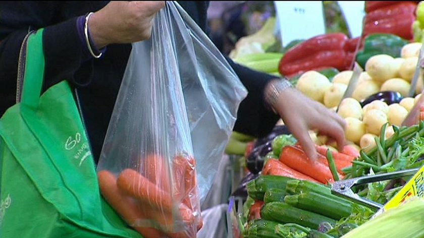 A shopper puts produce into a plastic bag while carrying a reusable bag over their other arm.