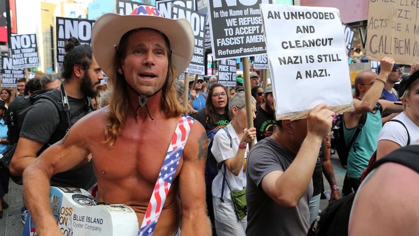 The Naked Cowboy stands next to a protester holding a sign that reads "An unhooded and clean-cut Nazi is still a Nazi".