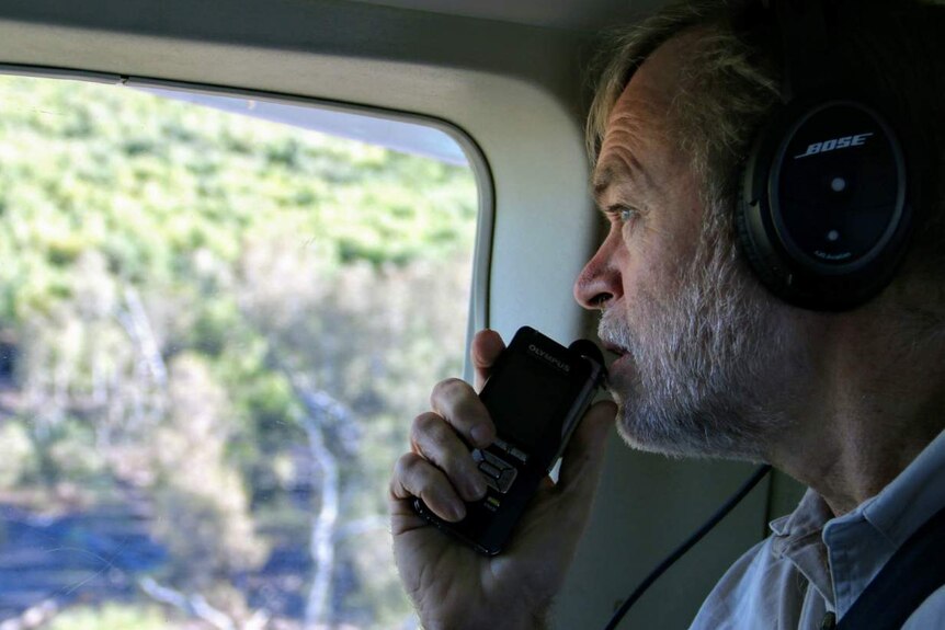 A grey bearded man in a plane talking into a receiver looking out a window