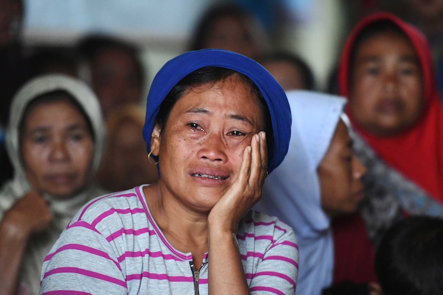 A young woman, wearing a blue hat and braces, holds her face as she cries with other women behind.