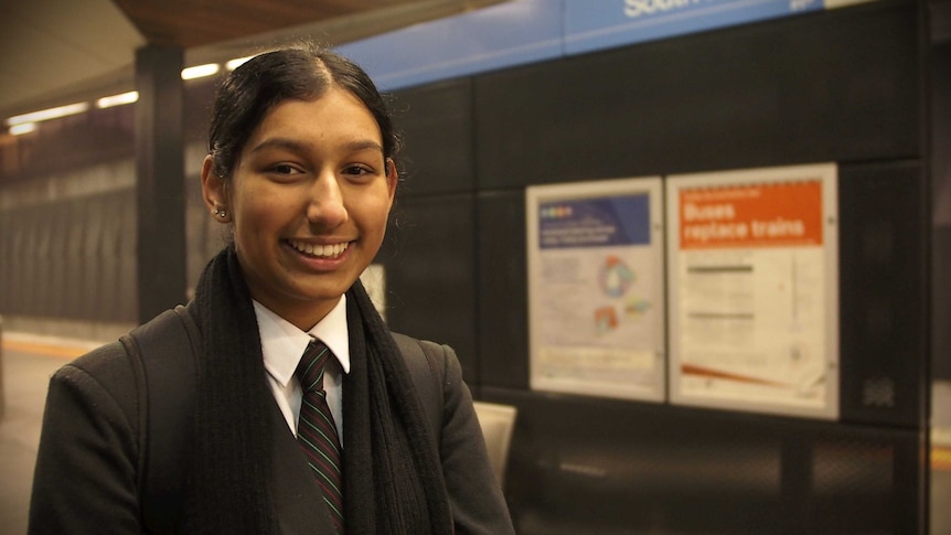 In a dimly-lit new train station platform, you view a young Australian student of South Asian descent in school uniform.