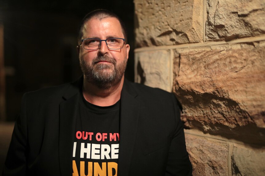 A man with glasses and a black jacket over a tshirt, leans against a sandstone wall. He has a neutral expression.
