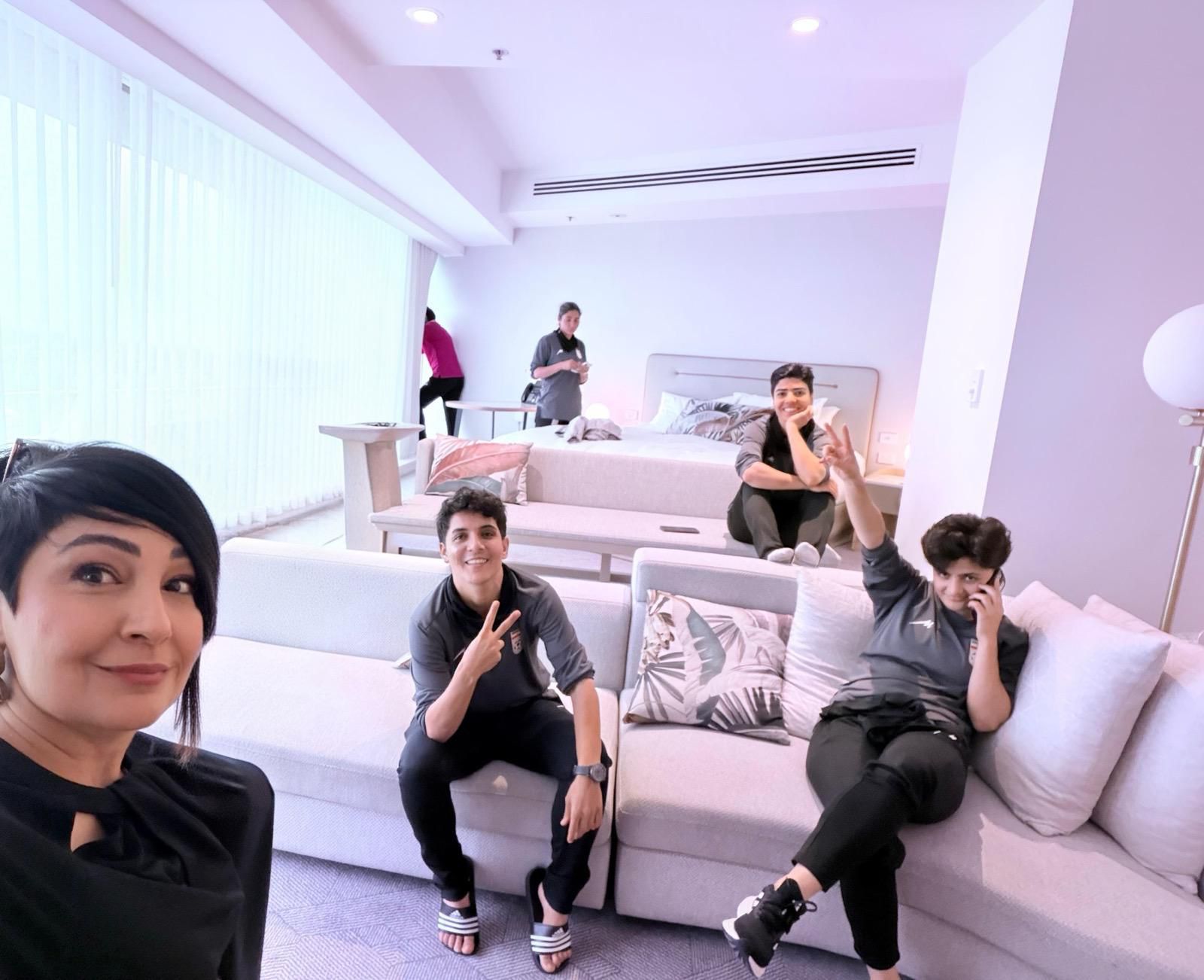A woman takes a selfie in a hotel room, with four other women in the background smiling