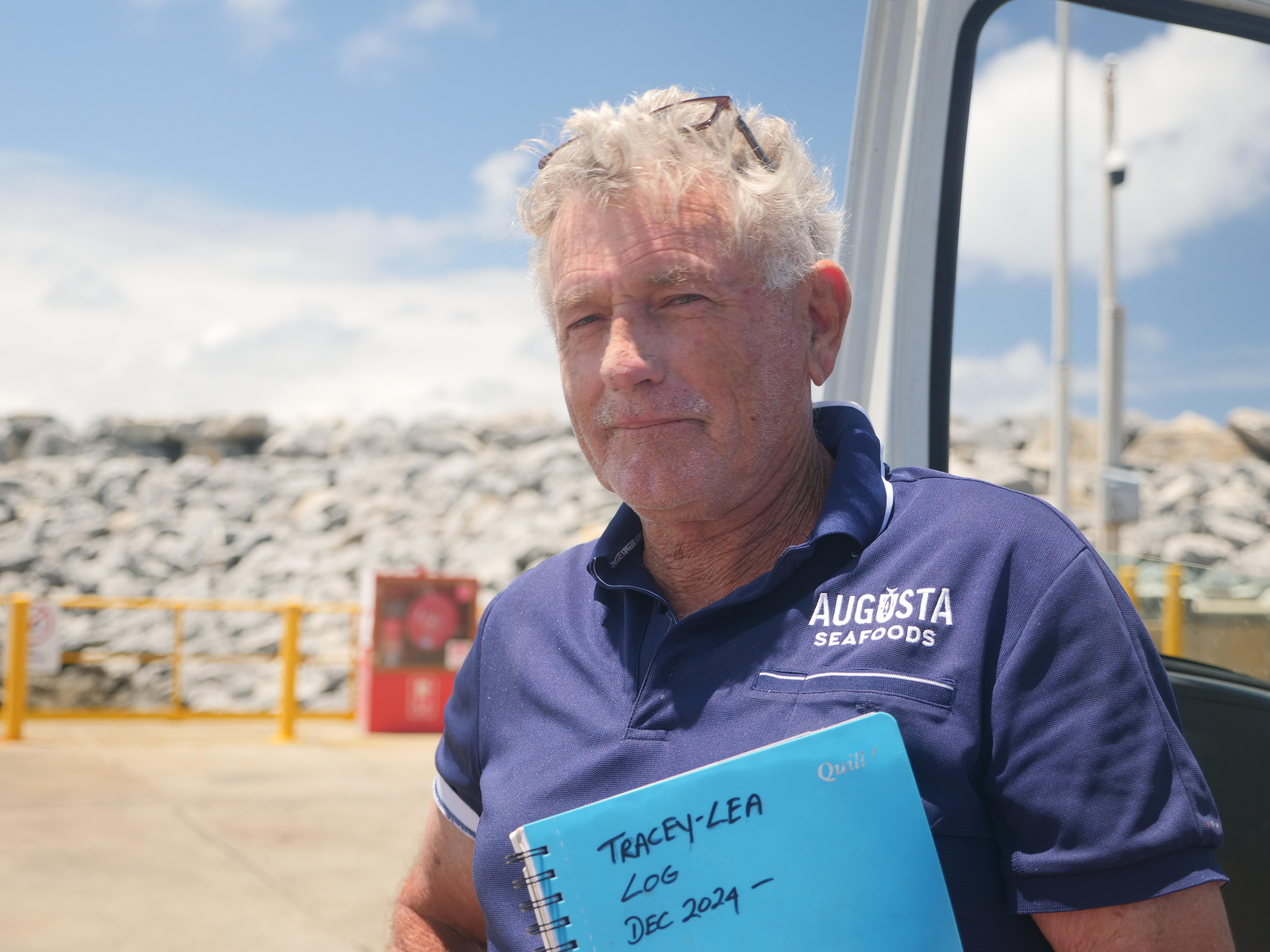 Un hombre de pelo blanco con un polo azul oscuro y mirando a la cámara con una libreta azul. 