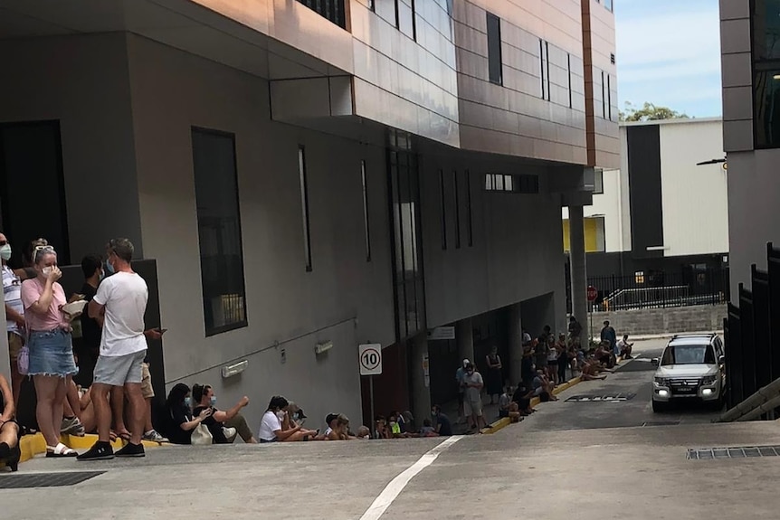 A long line of people snakes along a footpath alongside a building.